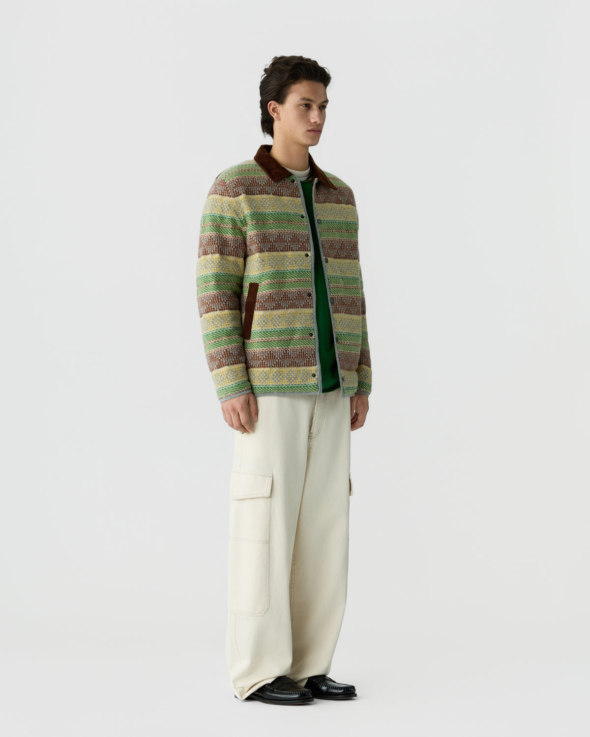 A young man stands against a plain background wearing a striped, multicolored knit jacket with a brown collar over a green shirt, paired with loose-fitting cream cargo pants and black shoes.