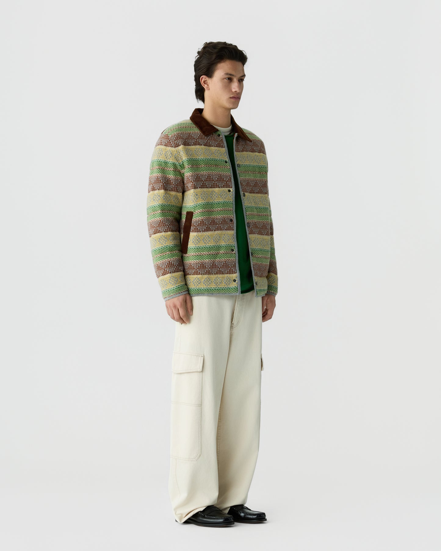 A young man stands against a plain background wearing a striped, multicolored knit jacket with a brown collar over a green shirt, paired with loose-fitting cream cargo pants and black shoes.