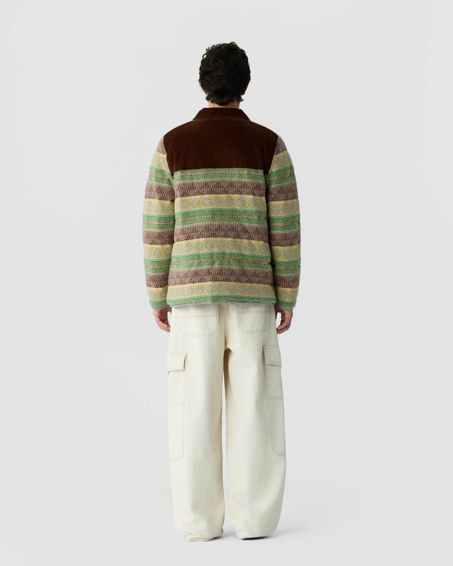 Man seen from behind wearing a multicolored striped fleece jacket with a brown collar and loose-fitting off-white cargo pants against a plain white background.