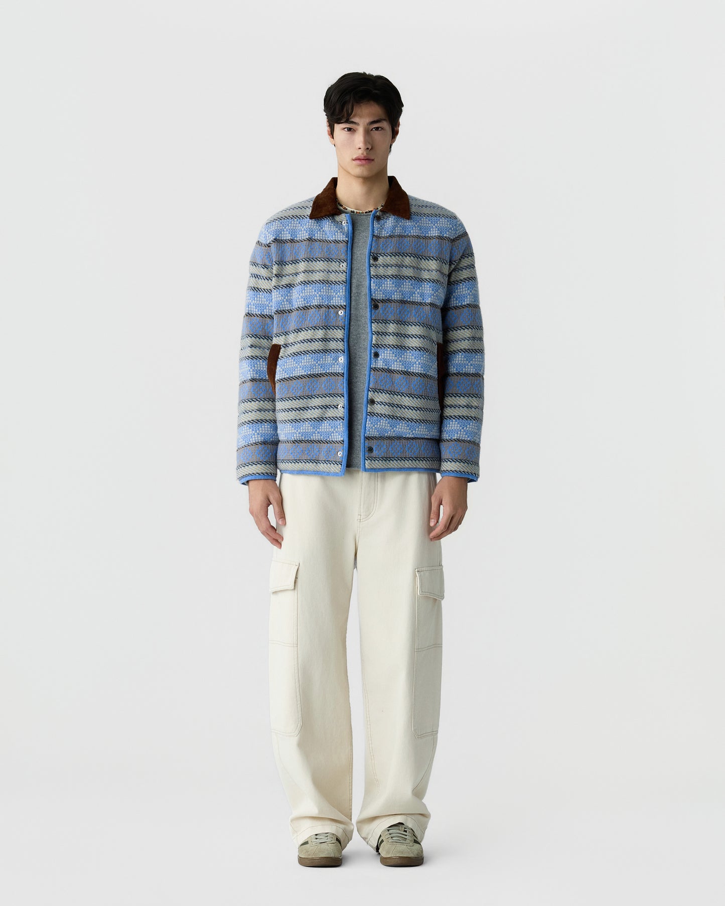 Young man wearing a blue and gray patterned jacket with a brown collar, light blue shirt, cream cargo pants, and beige sneakers, standing against a plain white background.