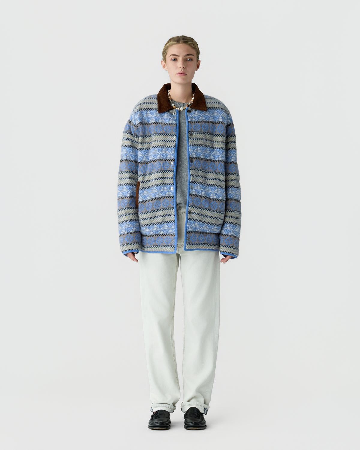 Young woman standing against a plain white background wearing a blue patterned oversized jacket with a brown collar, white pants, and black shoes.