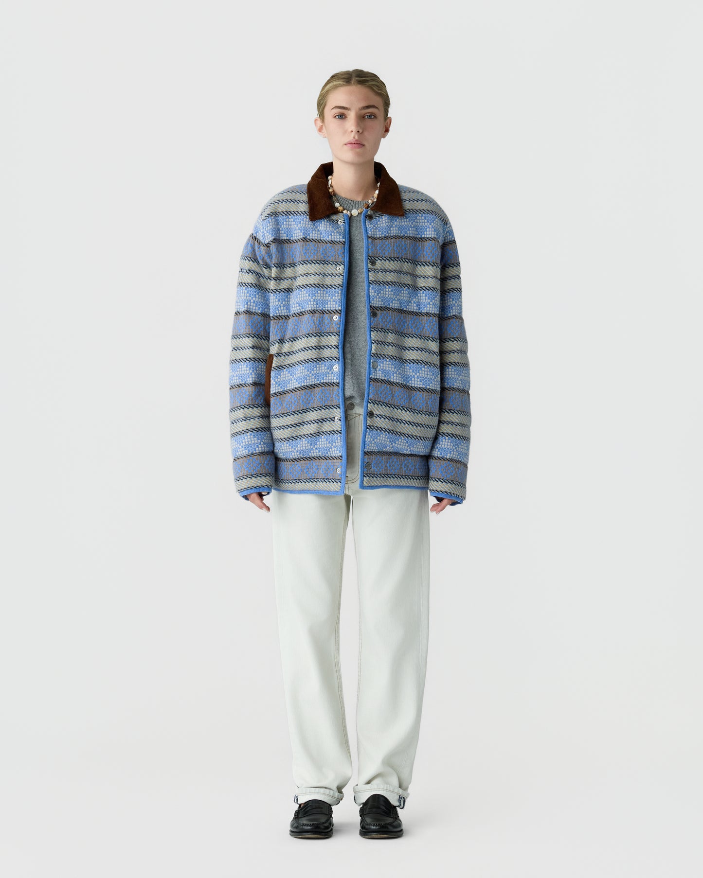 Young woman standing against a plain white background wearing a blue patterned oversized jacket with a brown collar, white pants, and black shoes.