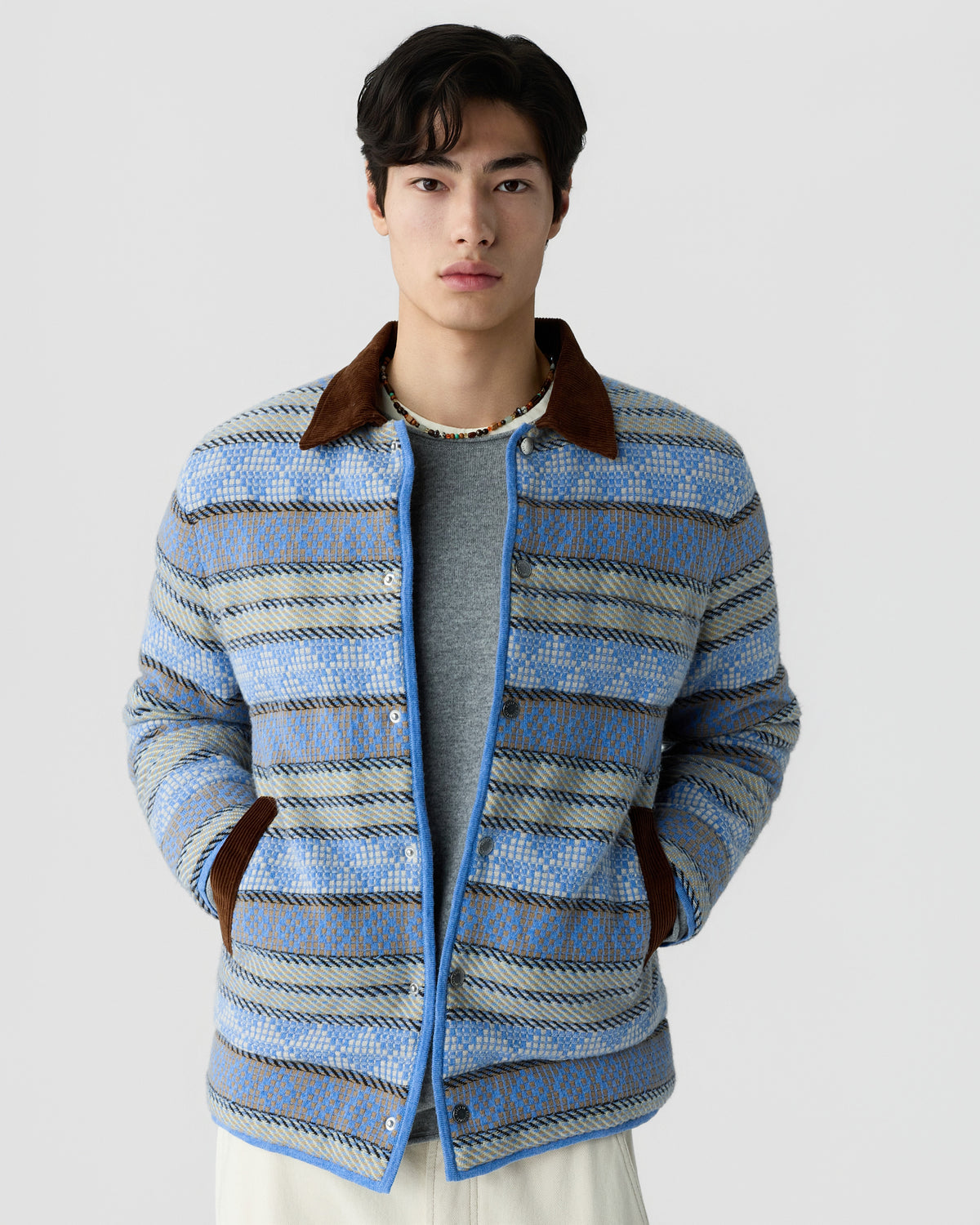 A young man wearing a blue and gray patterned jacket with a brown collar and cuffs, standing against a plain light background with hands in pockets.