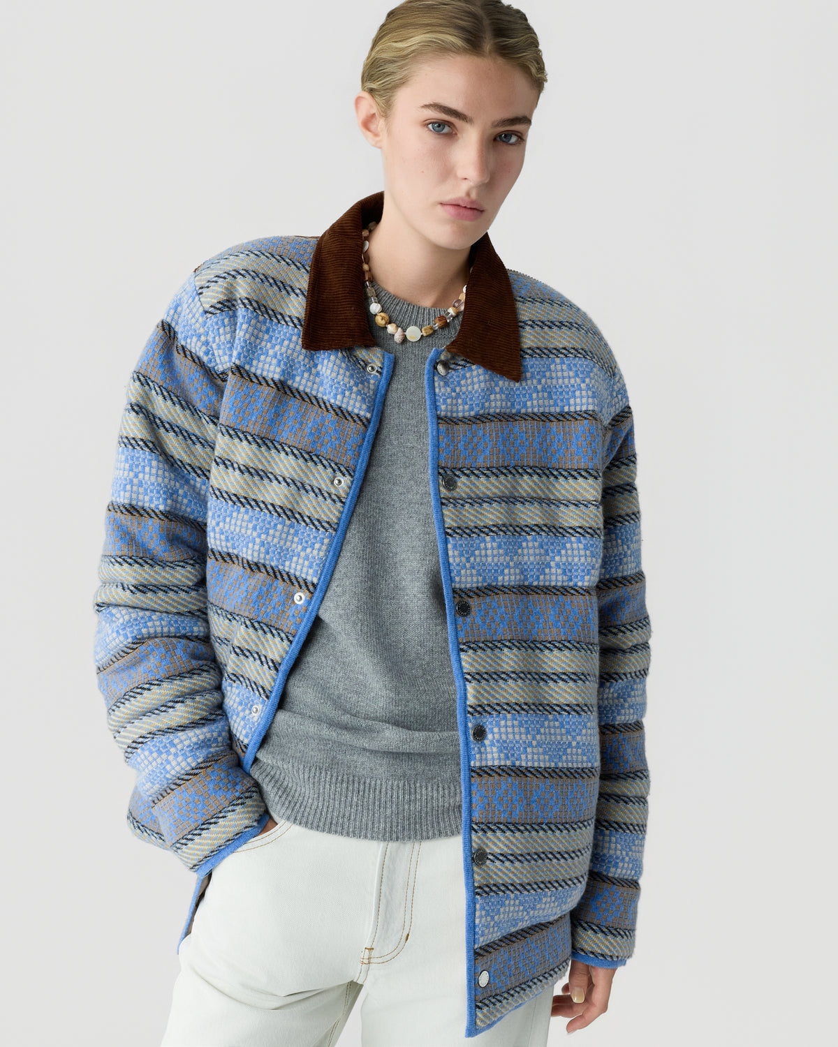 Woman wearing a blue and beige patterned quilted jacket with a brown collar over a gray sweater and white pants against a plain light background.