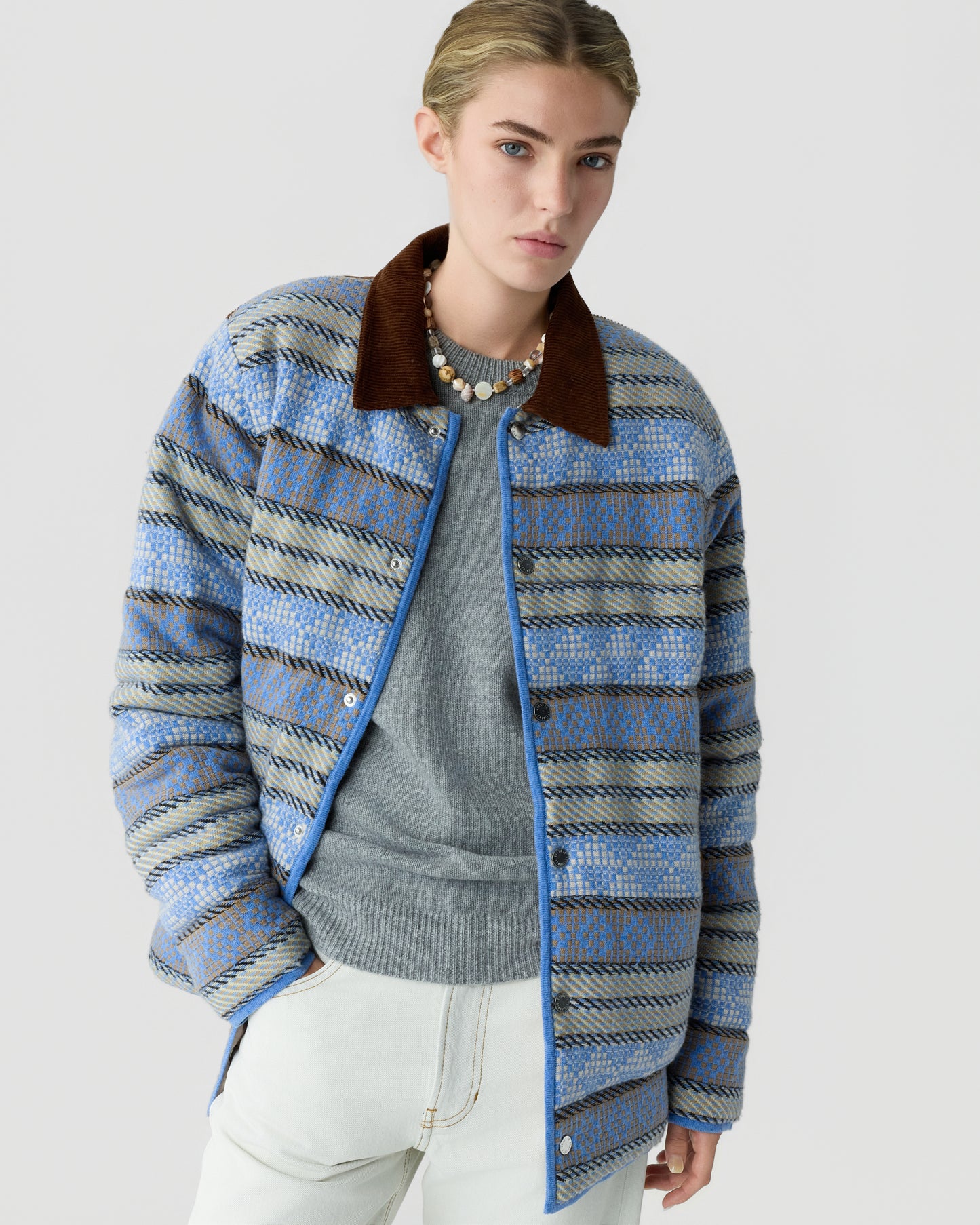 Woman wearing a blue and beige patterned quilted jacket with a brown collar over a gray sweater and white pants against a plain light background.