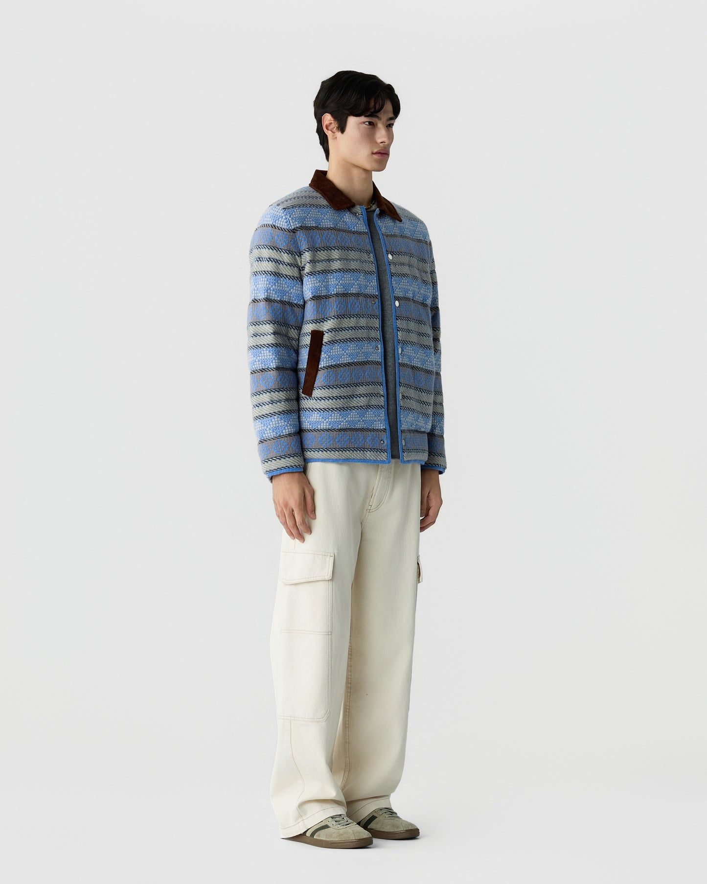 Young man wearing a blue and gray patterned jacket with brown accents over a light shirt, paired with loose white cargo pants and beige sneakers, standing against a plain white background.