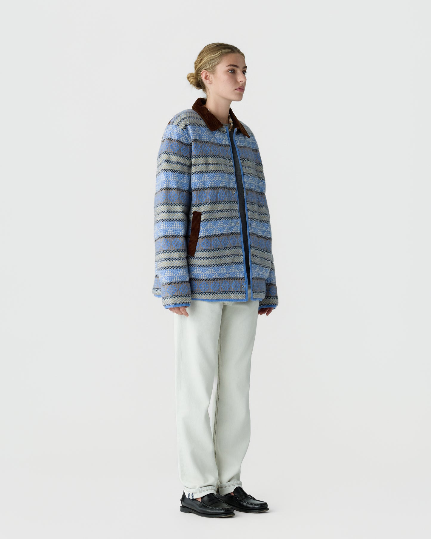 Young woman with blonde hair in a bun wearing a blue patterned jacket with brown collar and pockets, white pants, and black shoes, standing against a plain white background.