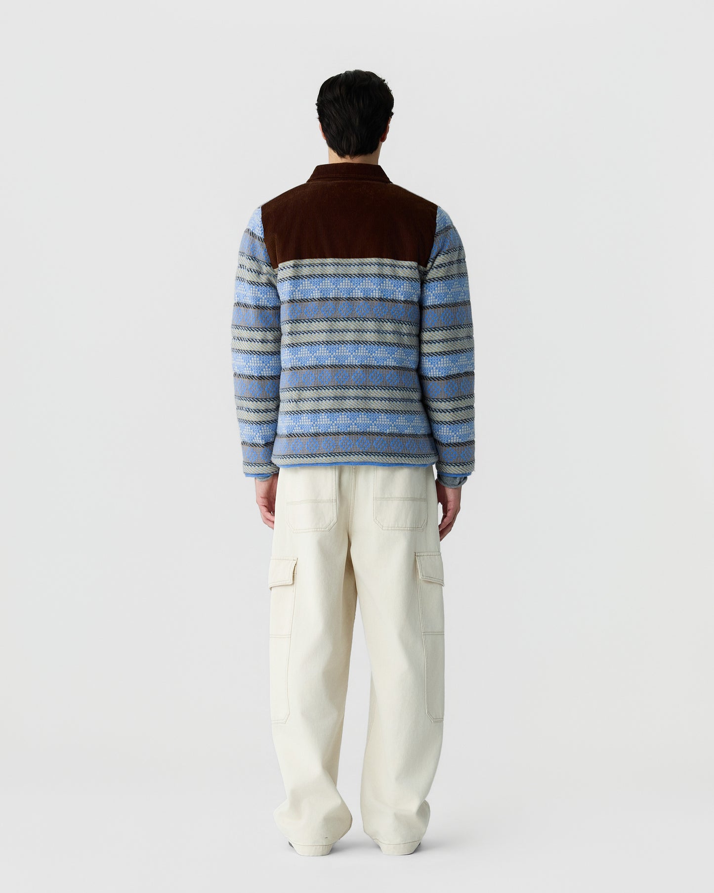 Man wearing a patterned blue and brown fleece jacket with cream-colored cargo pants standing facing away against a plain white background.