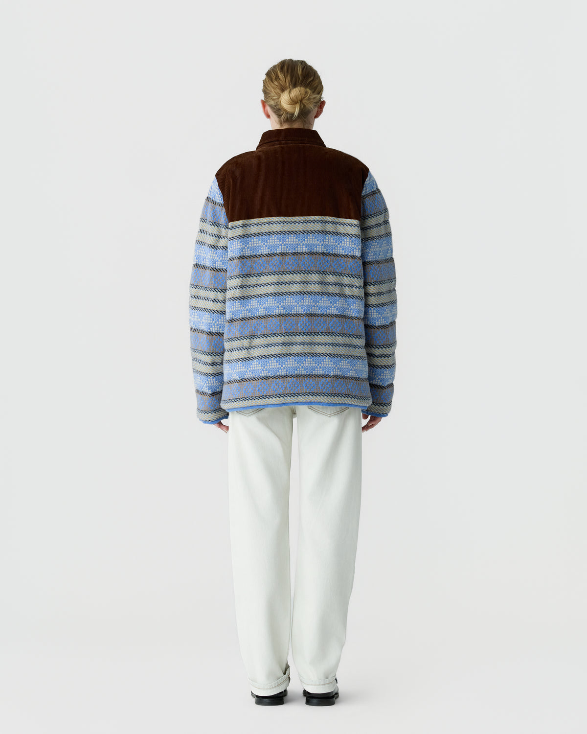 Person wearing a blue and brown patterned fleece jacket with a solid brown yoke, paired with white pants and black shoes, standing facing away against a plain light background.