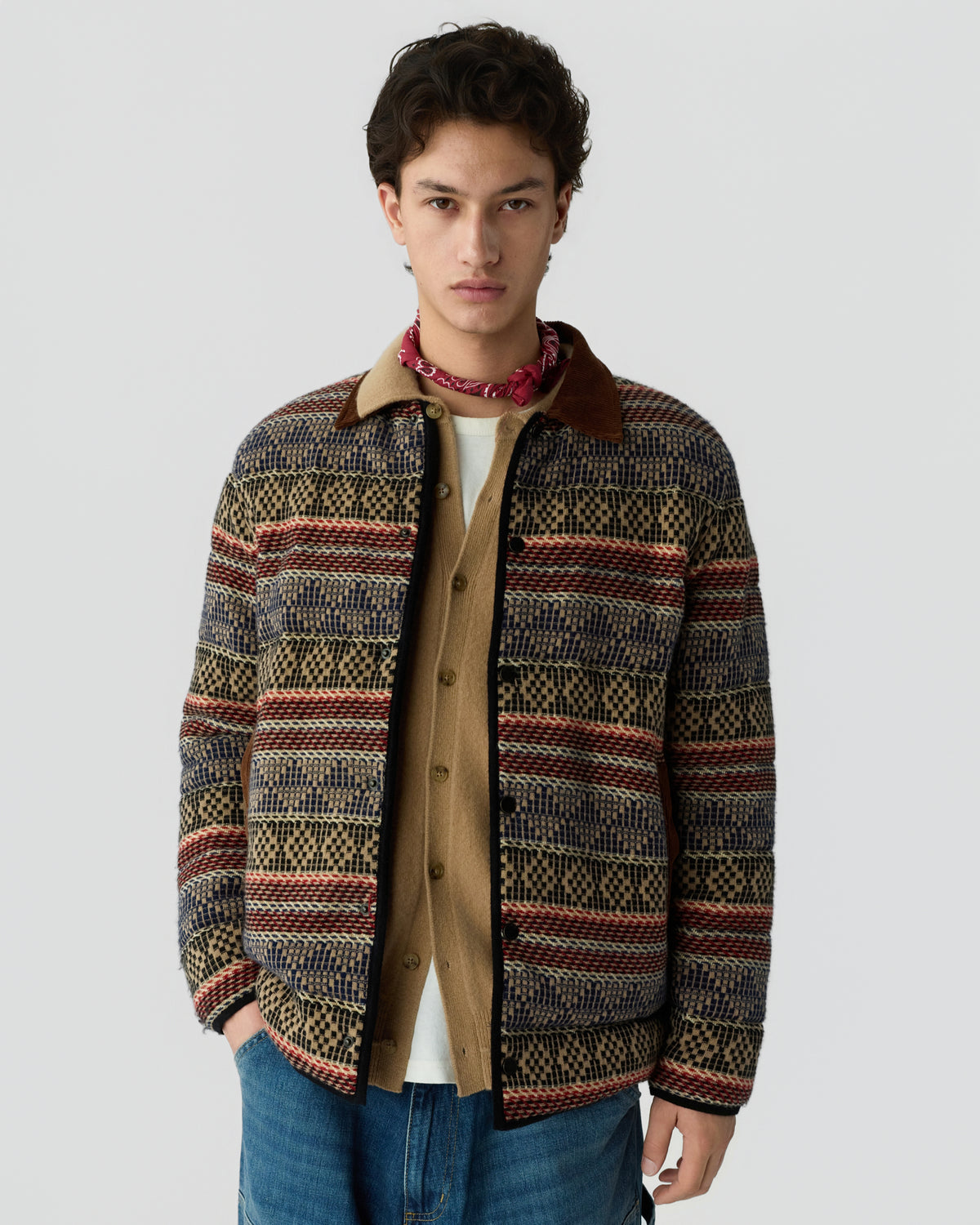 Young man standing against a light gray background wearing a patterned jacket over a beige shirt with a red neck scarf and blue jeans
