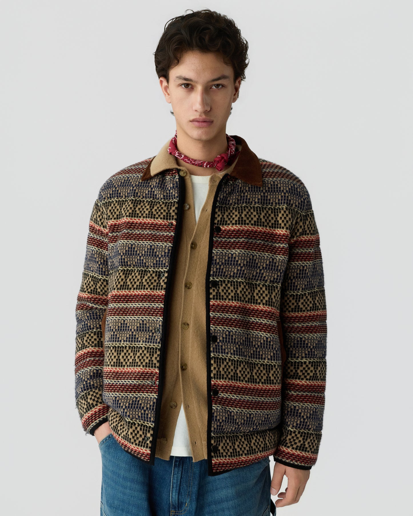 Young man standing against a light gray background wearing a patterned jacket over a beige shirt with a red neck scarf and blue jeans