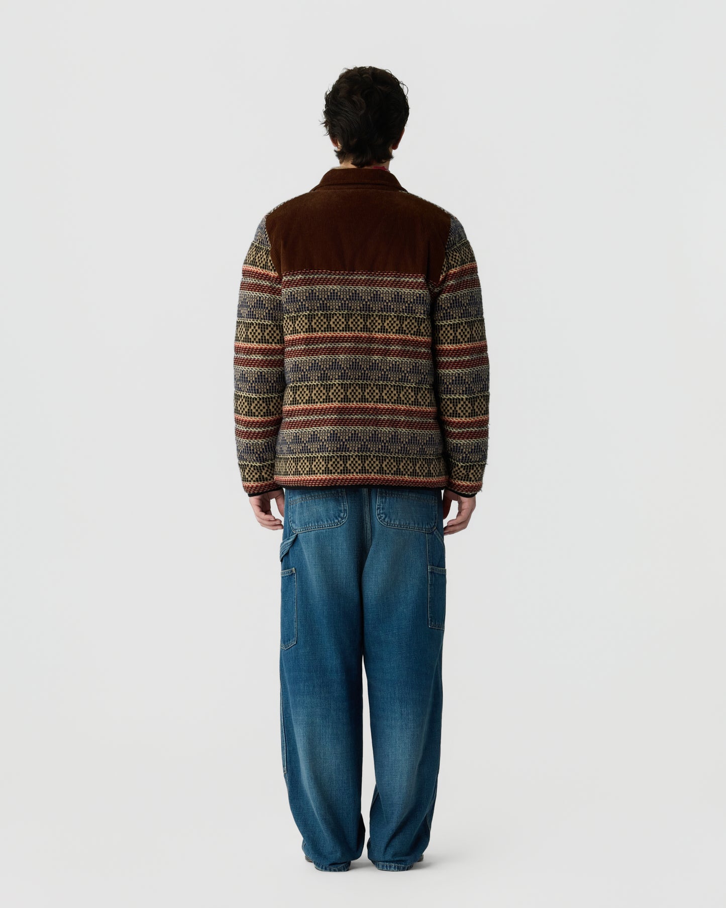 Back view of a person wearing a brown patterned knit sweater with multicolored horizontal bands and loose blue jeans against a light gray background
