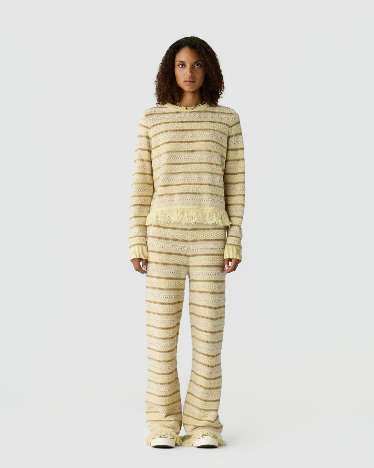 Model wearing a cream and beige striped knit sweater with a fringed hem and matching flared pants against a light gray studio background