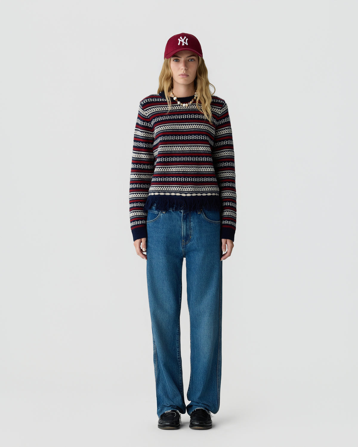 Woman wearing a striped knit sweater with blue wide leg jeans black shoes and a maroon baseball cap against a plain light gray studio background