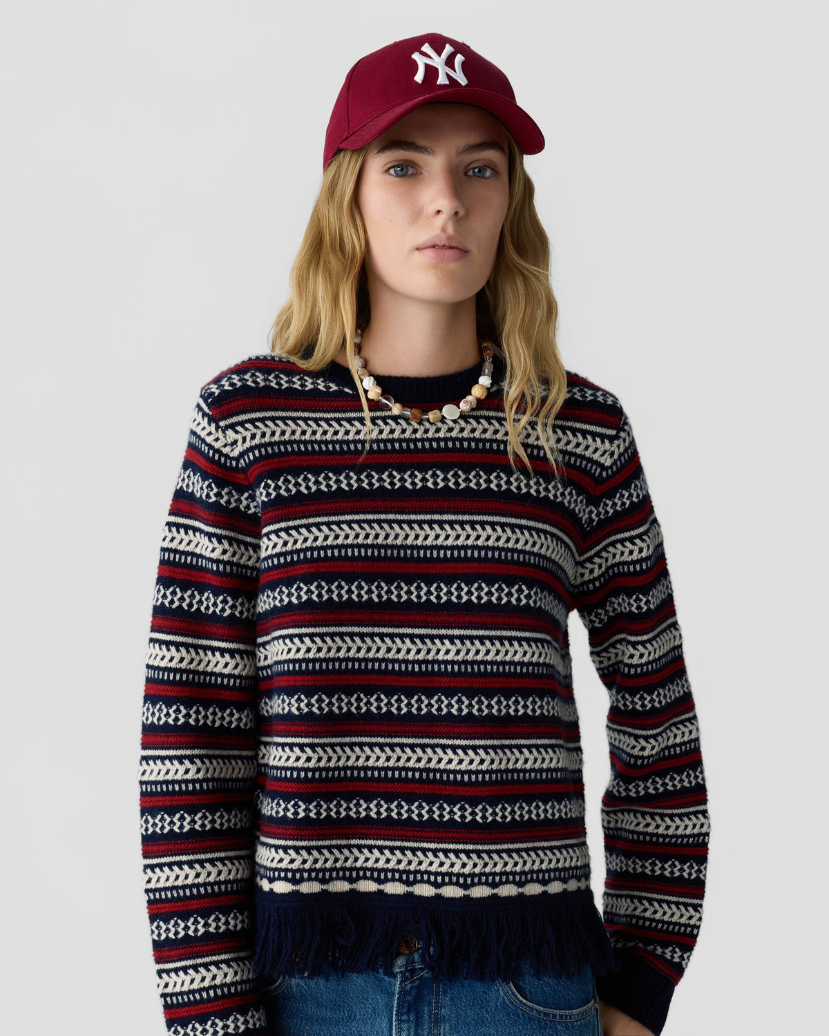 Woman wearing a navy patterned knit sweater with fringe hem, layered necklaces, blue jeans, and a burgundy baseball cap against a light gray background