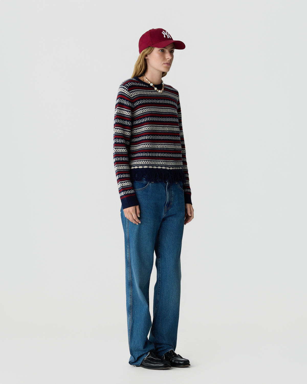 Model wearing a striped knit sweater with blue jeans and a burgundy baseball cap while standing against a light gray studio background