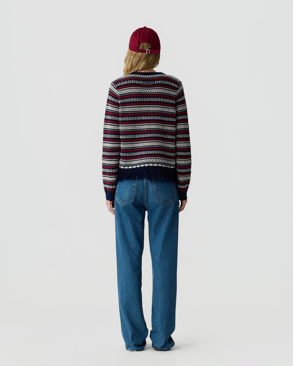 Person seen from behind wearing a patterned knit sweater with blue jeans and a burgundy cap against a light gray background