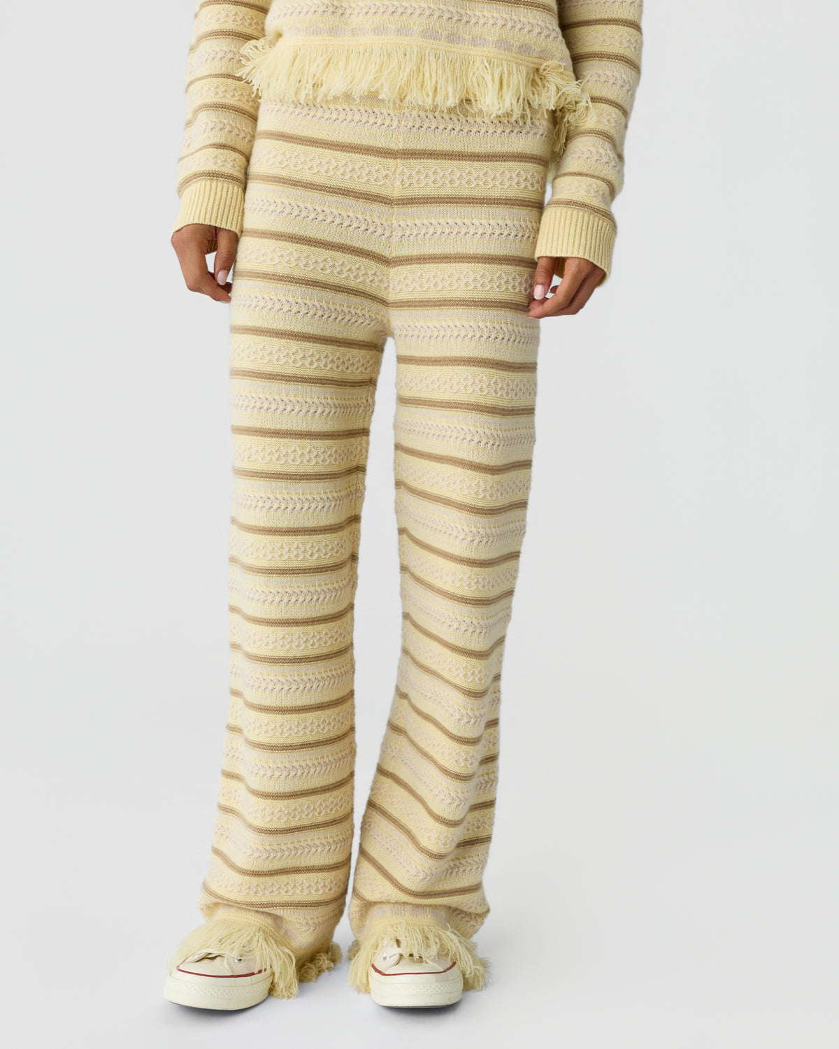 Cropped view of a person wearing a cream textured knit sweater and matching flared trousers with horizontal stripes and cream sneakers with fringe details