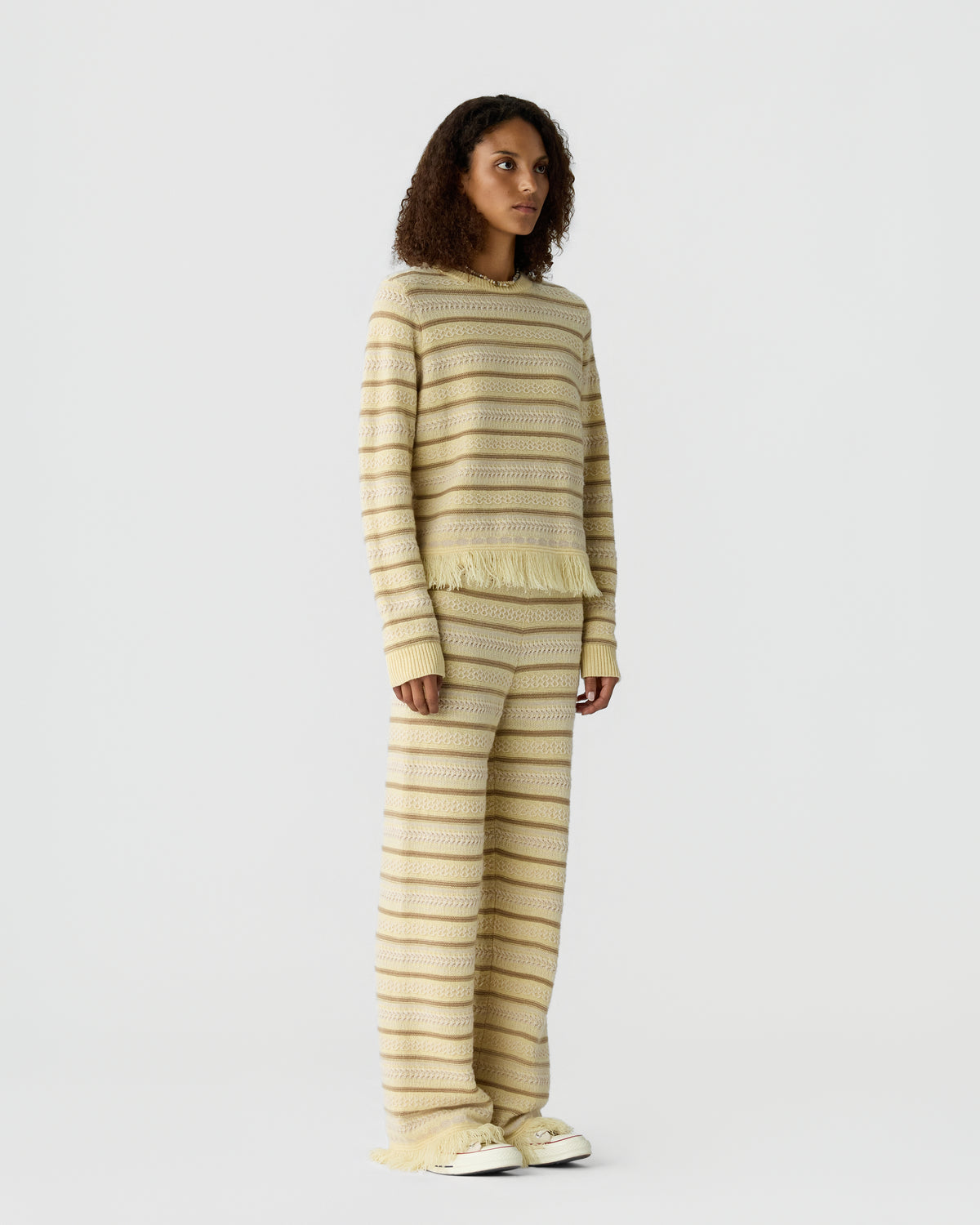 Model wearing a cream striped knit sweater and matching wide leg trousers with frayed hems and white sneakers against a light gray background