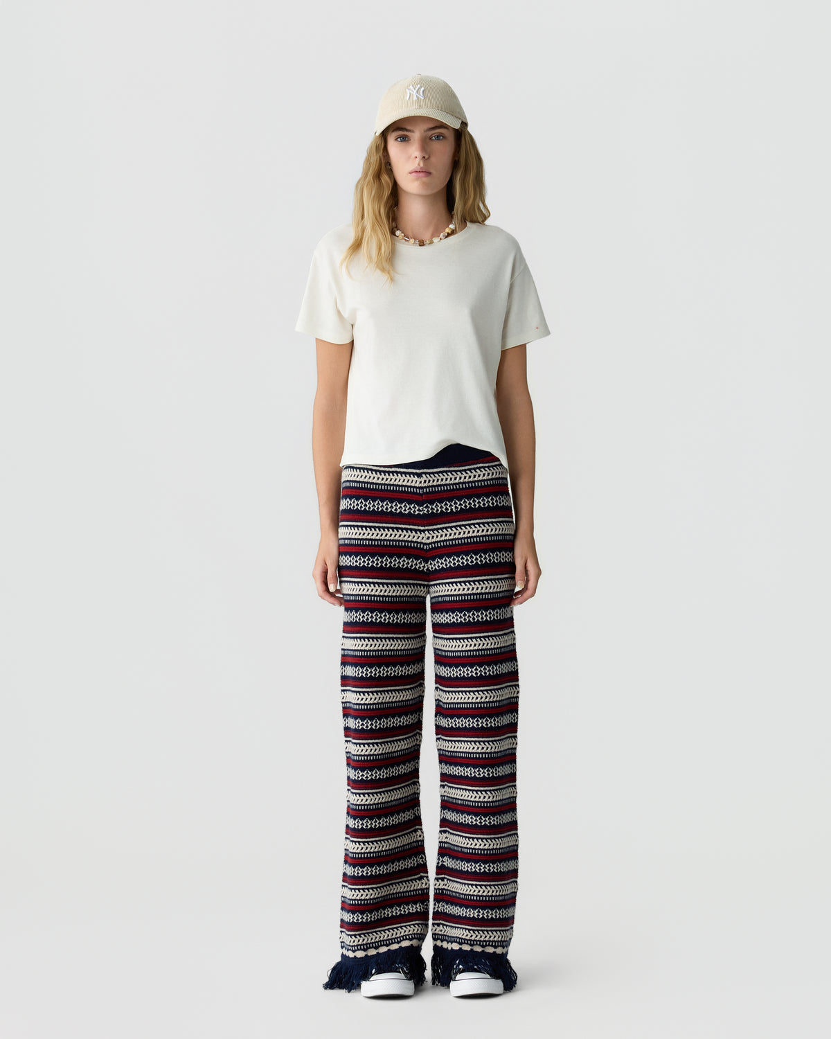 Woman wearing a white T shirt, beige baseball cap, patterned wide leg trousers, and white sneakers against a light gray studio background