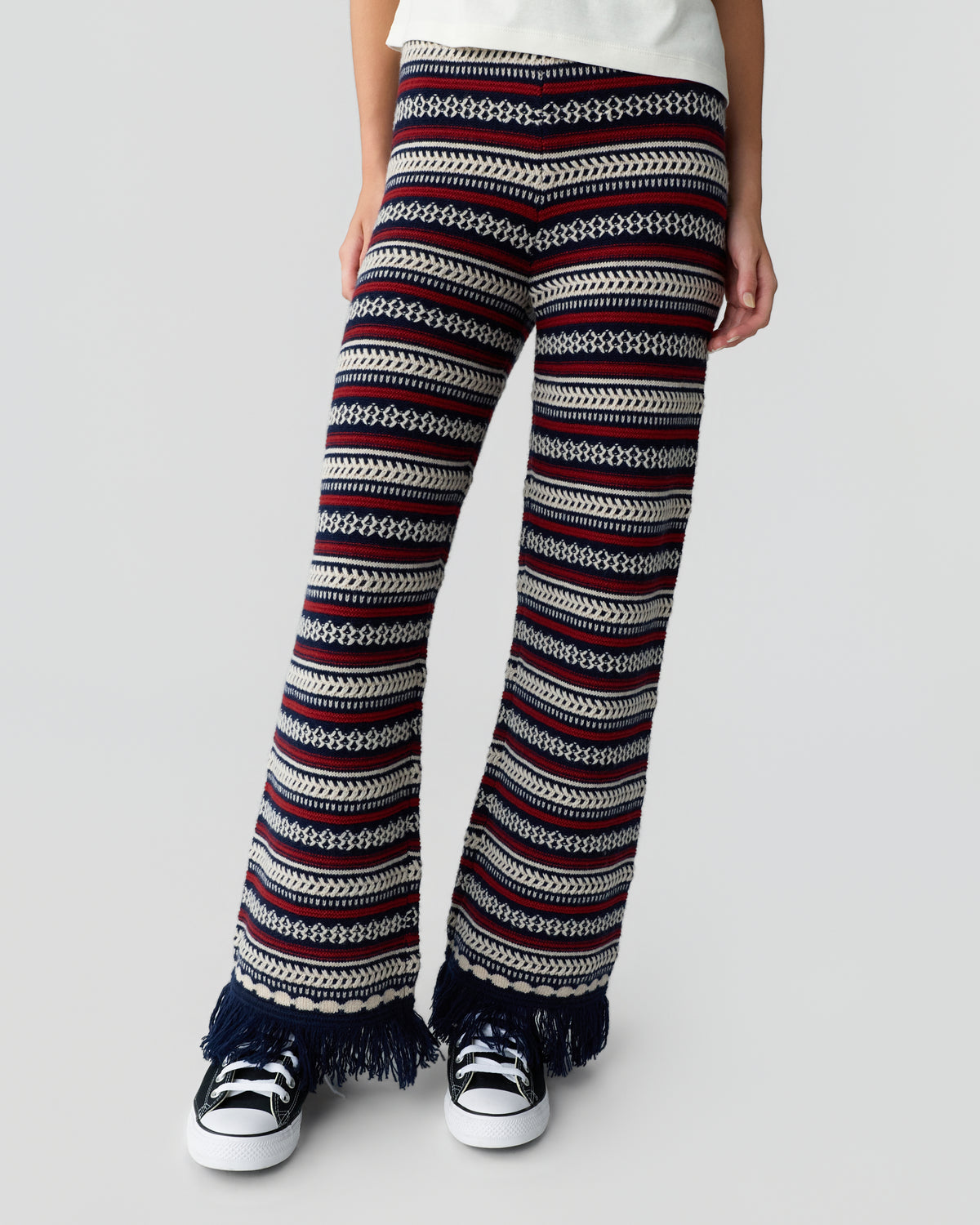 Person wearing patterned striped knit trousers with fringed hems and black and white sneakers against a light gray background