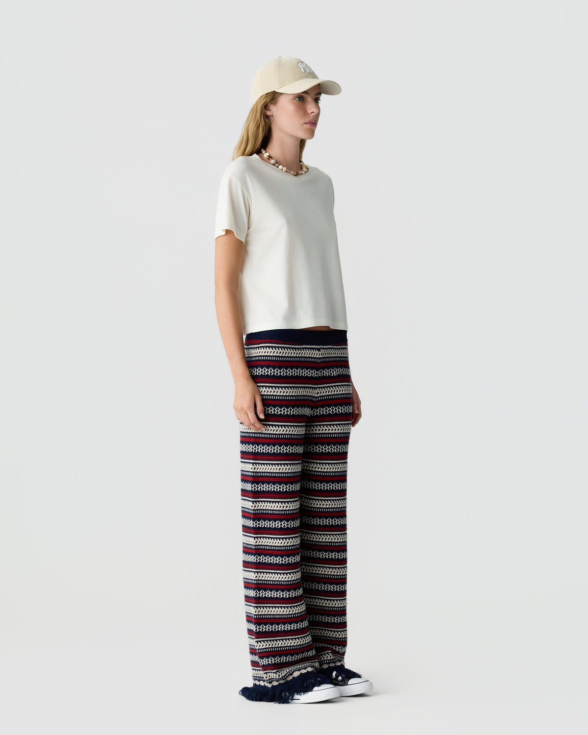 Woman wearing a white T-shirt, beige cap, patterned wide-leg trousers, and white sneakers against a light gray studio background.