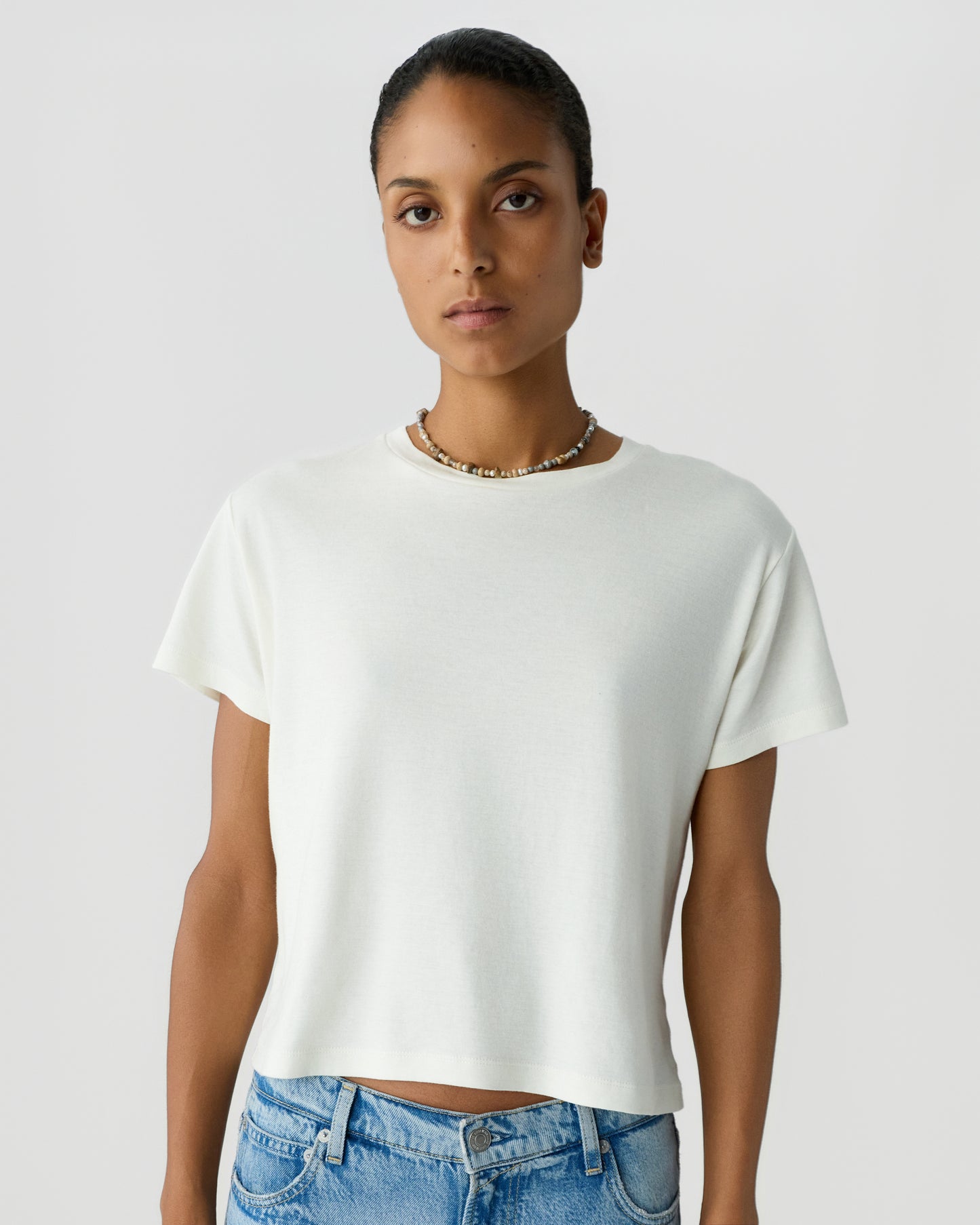 Woman with short dark hair wearing a plain white short sleeve T shirt and light blue jeans against a pale gray studio background