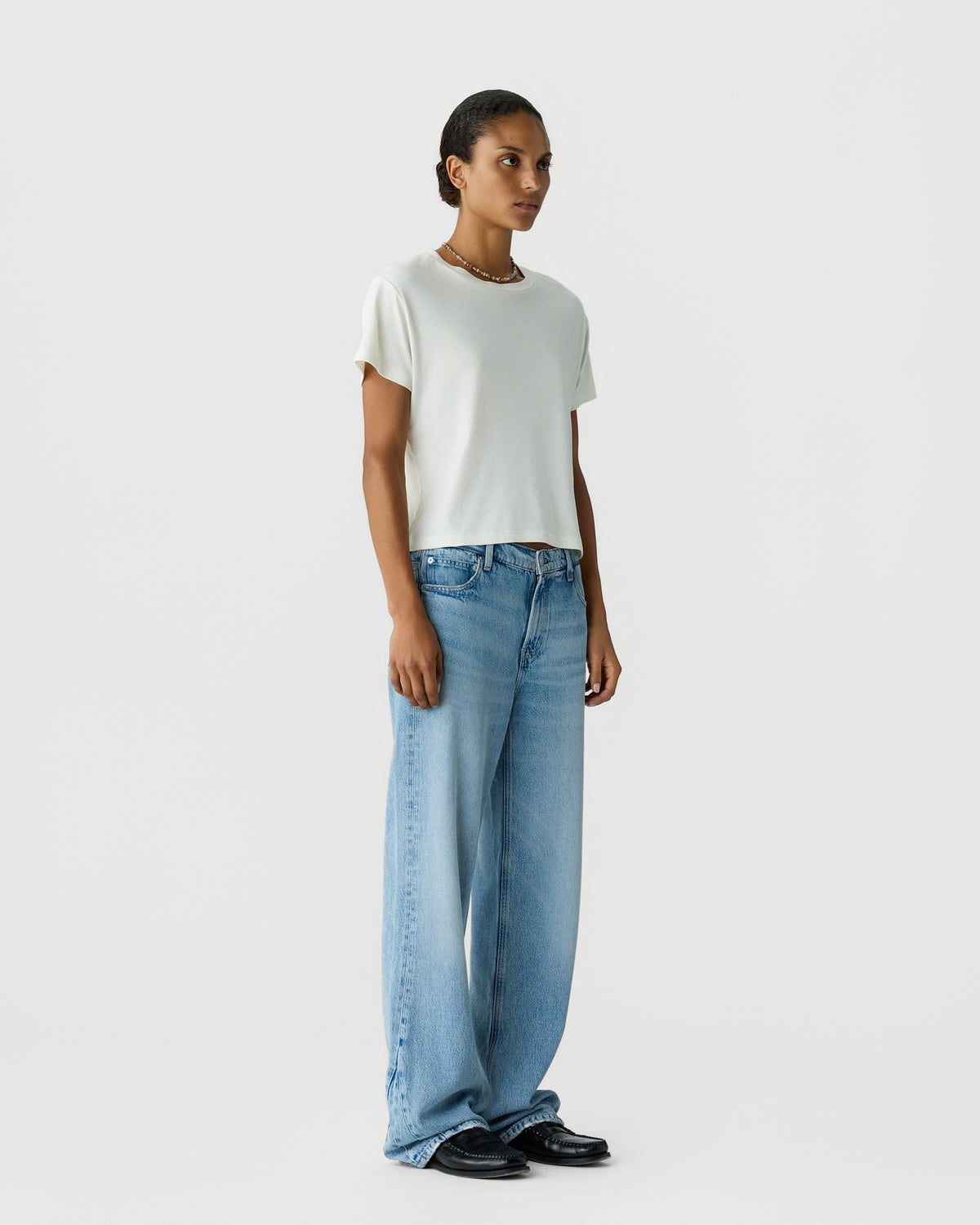 Model wearing a white short sleeve shirt with wide leg blue jeans and black shoes against a light gray studio background