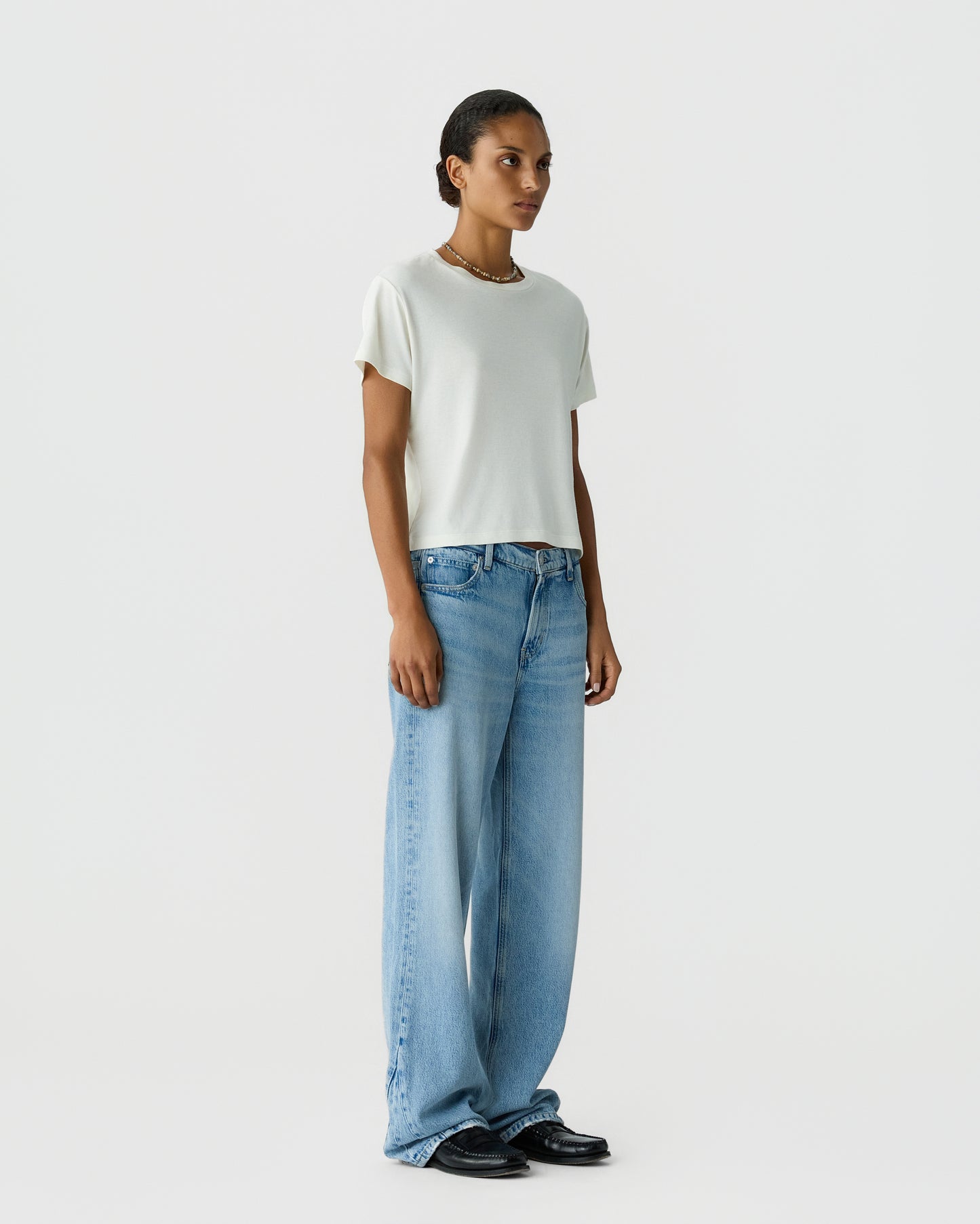 Model wearing a white short sleeve shirt with wide leg blue jeans and black shoes against a light gray studio background
