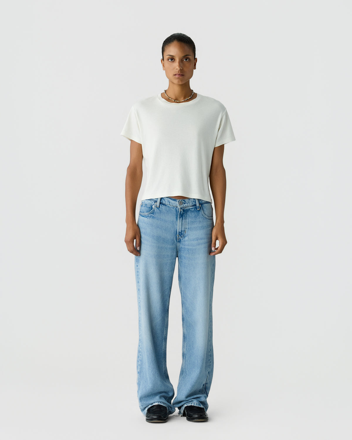 Person standing against a light gray background wearing a plain white T-shirt, loose blue jeans, black shoes, and a necklace.