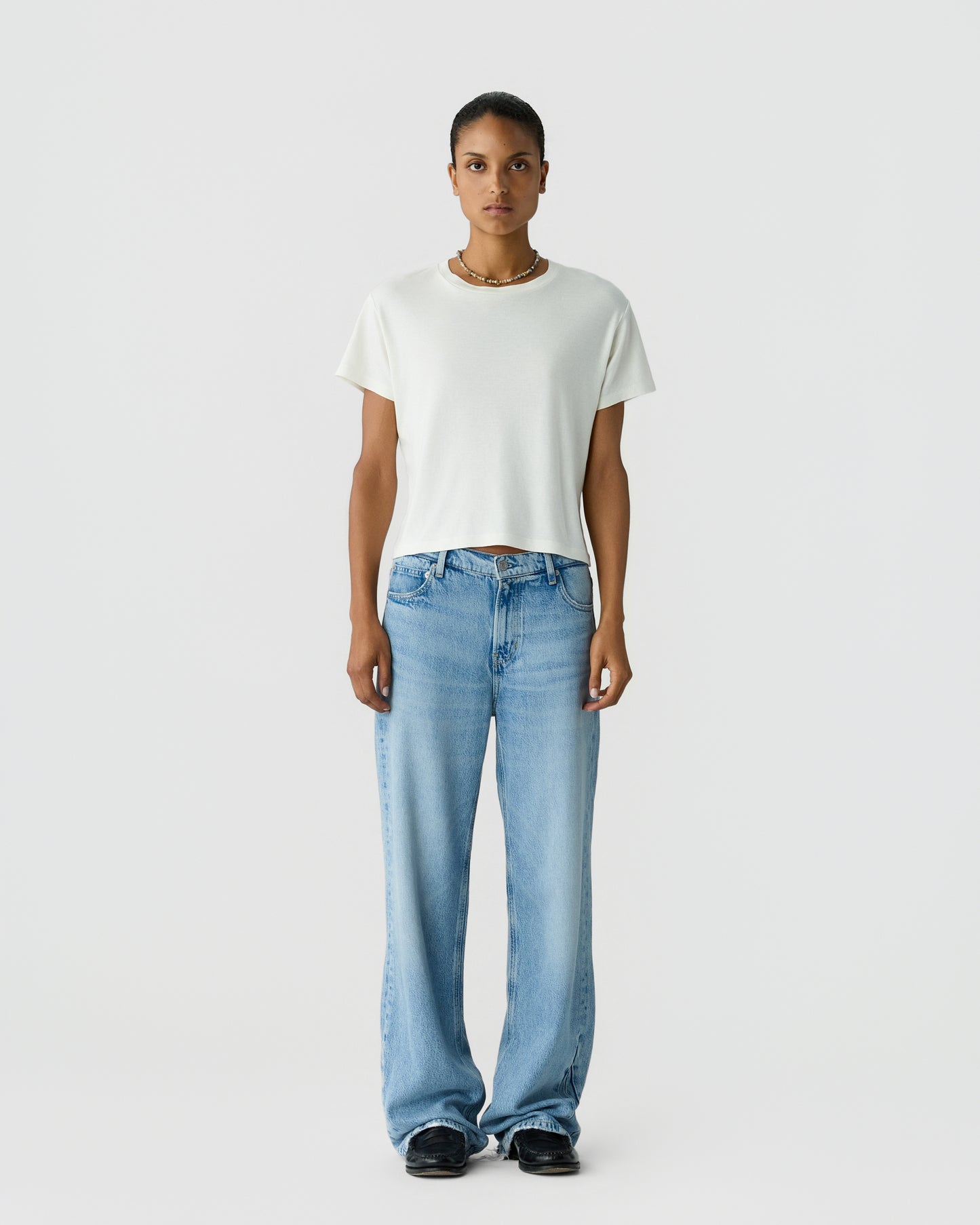 Person standing against a light gray background wearing a plain white T-shirt, loose blue jeans, black shoes, and a necklace.