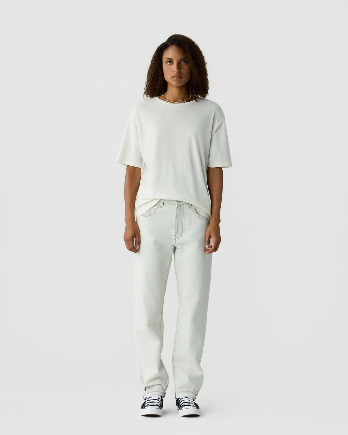 Woman with curly hair standing against a light gray background, wearing a white T shirt, light trousers, and black and white sneakers.