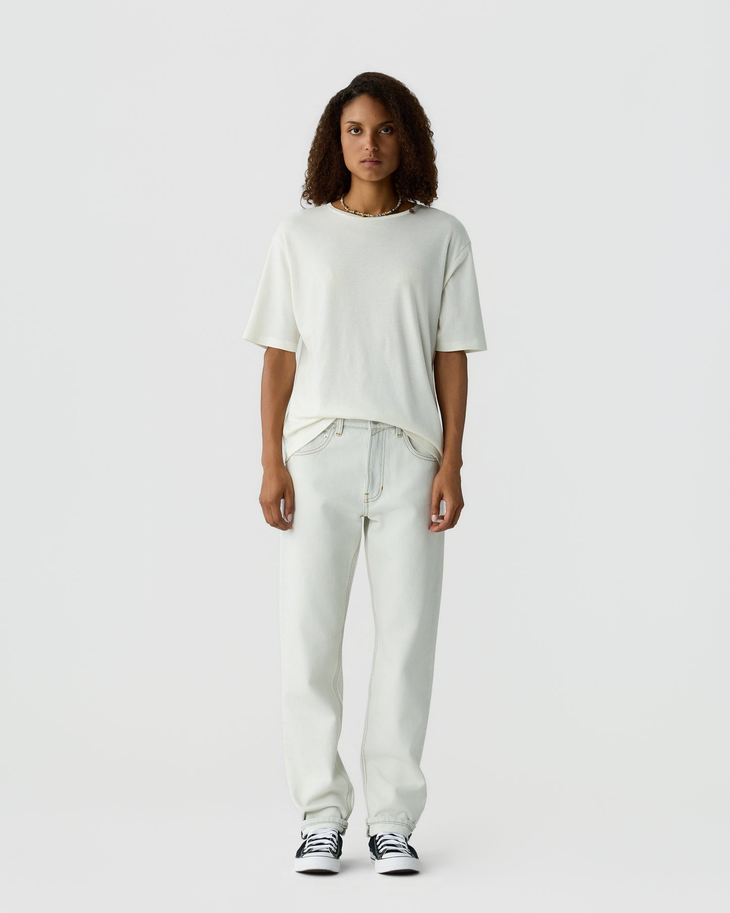 Woman with curly hair standing against a light gray background, wearing a white T shirt, light trousers, and black and white sneakers.
