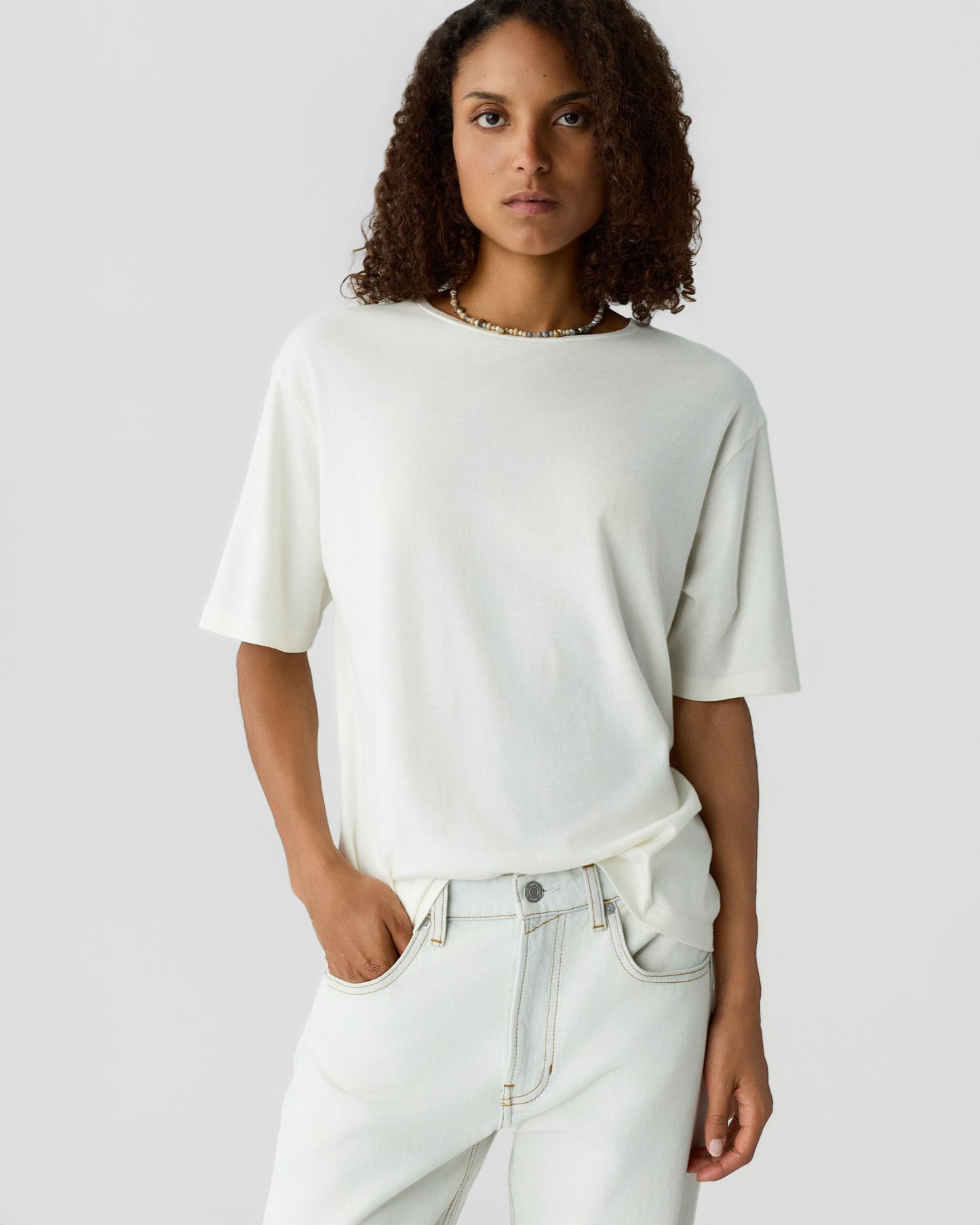 Woman with curly hair wearing a loose white short sleeve shirt and high waisted white jeans with hands in pockets against a light gray background
