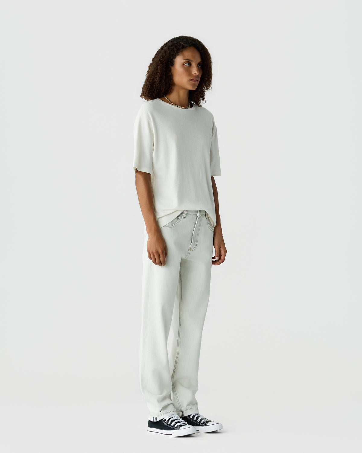 Model with curly hair wearing an off white short sleeve top and relaxed matching trousers with black and white sneakers against a light gray background