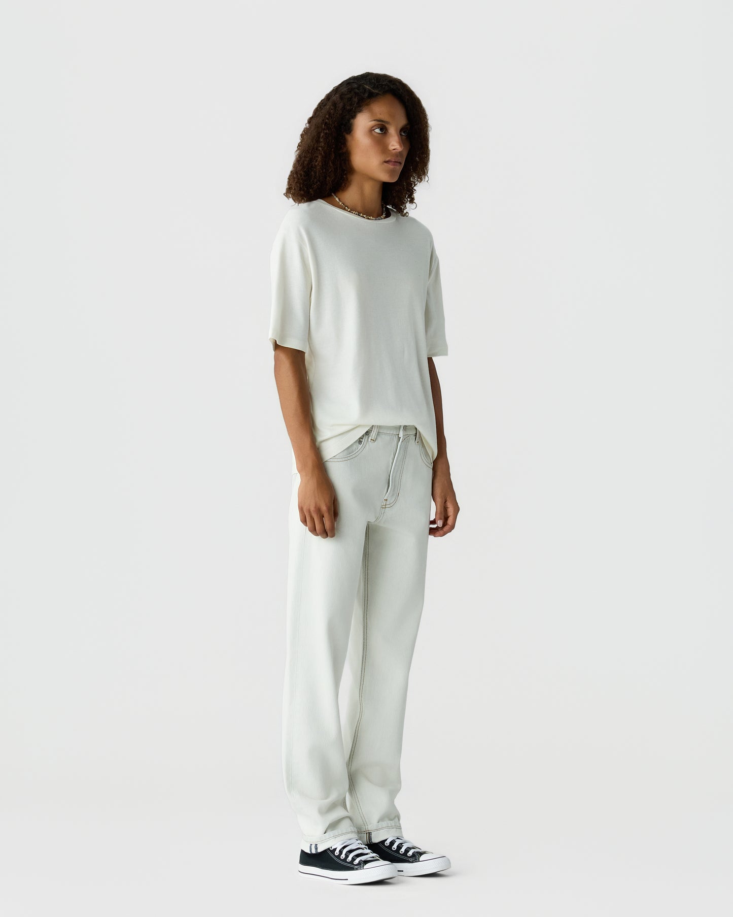 Model with curly hair wearing an off white short sleeve top and relaxed matching trousers with black and white sneakers against a light gray background
