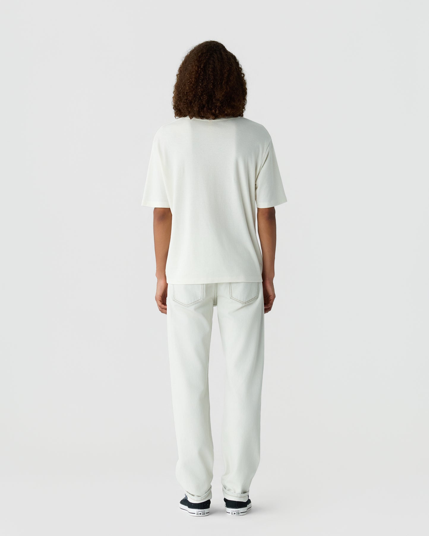 Rear view of a person with curly hair wearing a white short sleeve shirt and loose white pants with sneakers against a light gray studio background