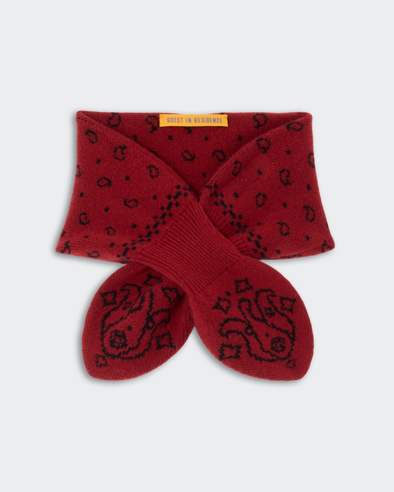 Red knitted scarf with black paisley and dot patterns, featuring a ribbed section and a yellow label with text at the top center.