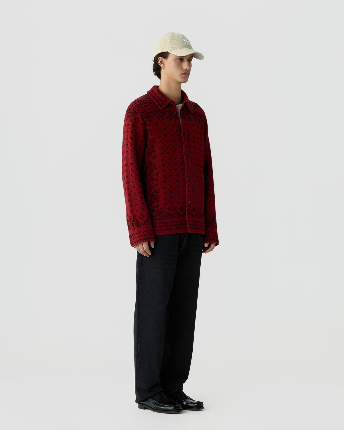 A young man wearing a beige baseball cap, a red patterned long-sleeve knit jacket, loose black pants, and black shoes standing against a plain white background.