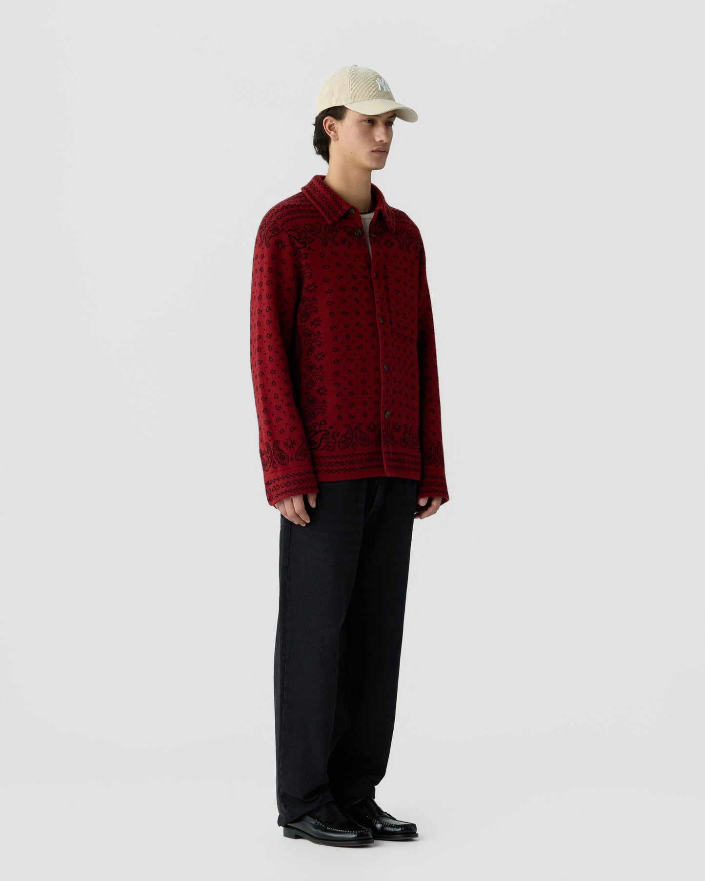 A young man wearing a beige baseball cap, a red patterned long-sleeve knit jacket, loose black pants, and black shoes standing against a plain white background.