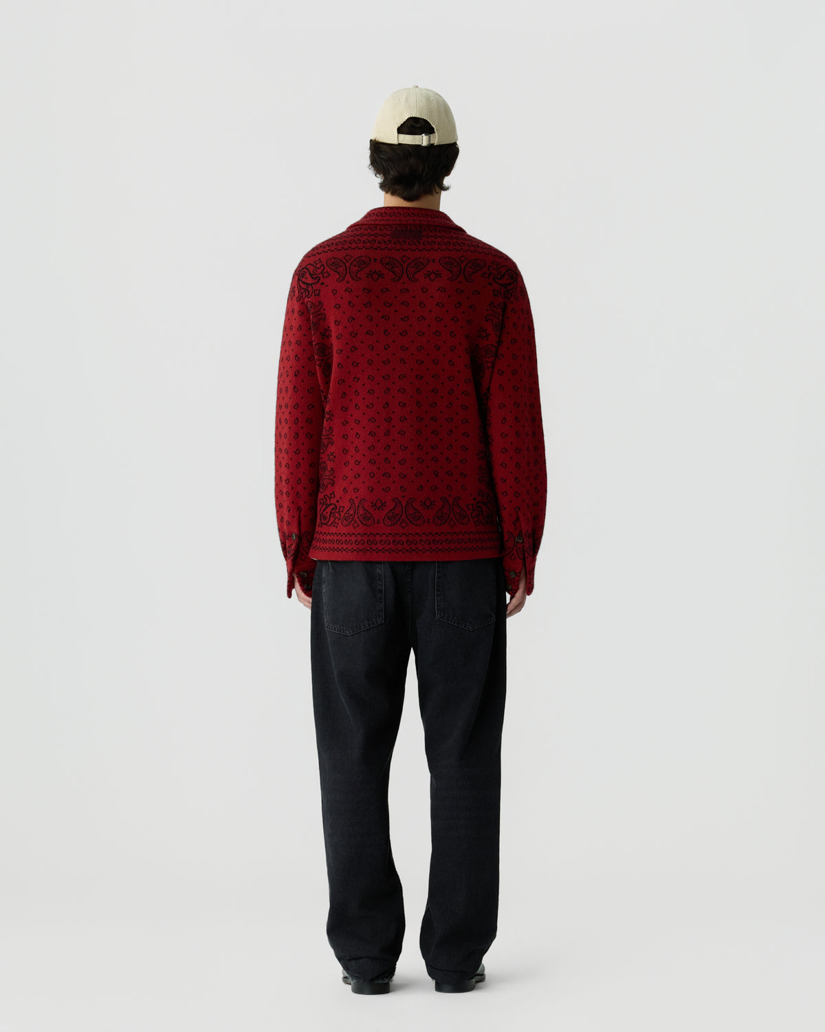 Man wearing a red patterned long-sleeve shirt, black pants, and a white baseball cap, standing facing away against a plain light background