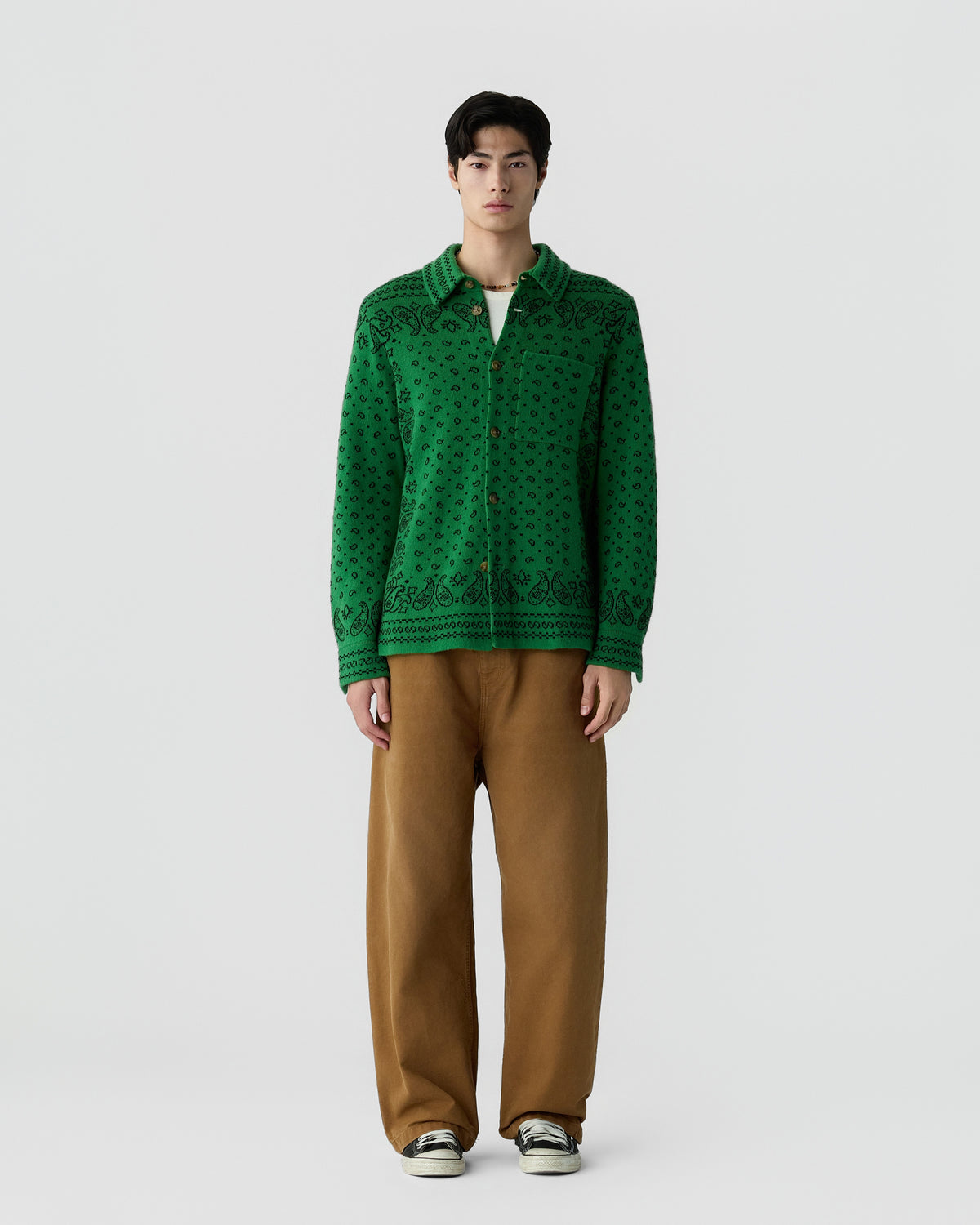 A man stands facing forward wearing a green patterned button-up cardigan, brown loose-fitting pants, and black and white sneakers against a plain light gray background.