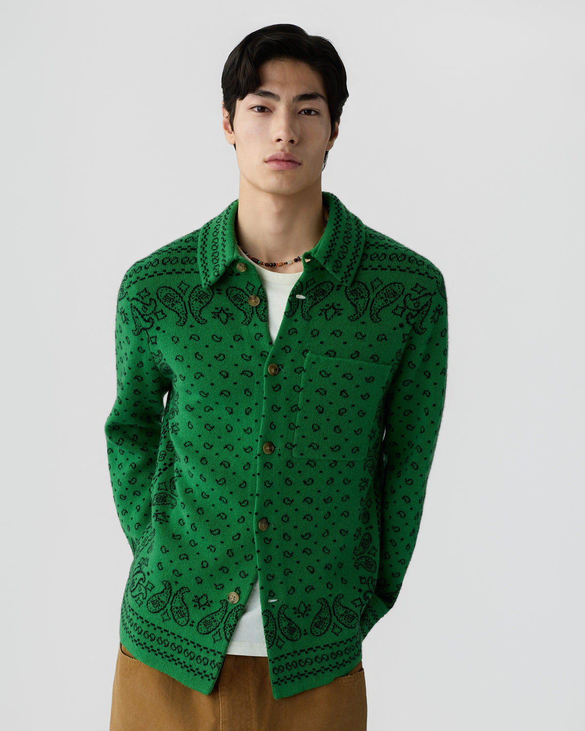 Young man wearing a green button-up cardigan with black paisley patterns over a white shirt, standing against a plain light gray background.