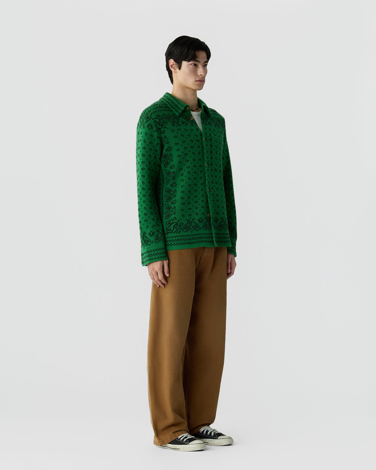 Young man wearing a green patterned long-sleeve shirt, brown wide-leg pants, and black and white sneakers standing against a plain light gray background.