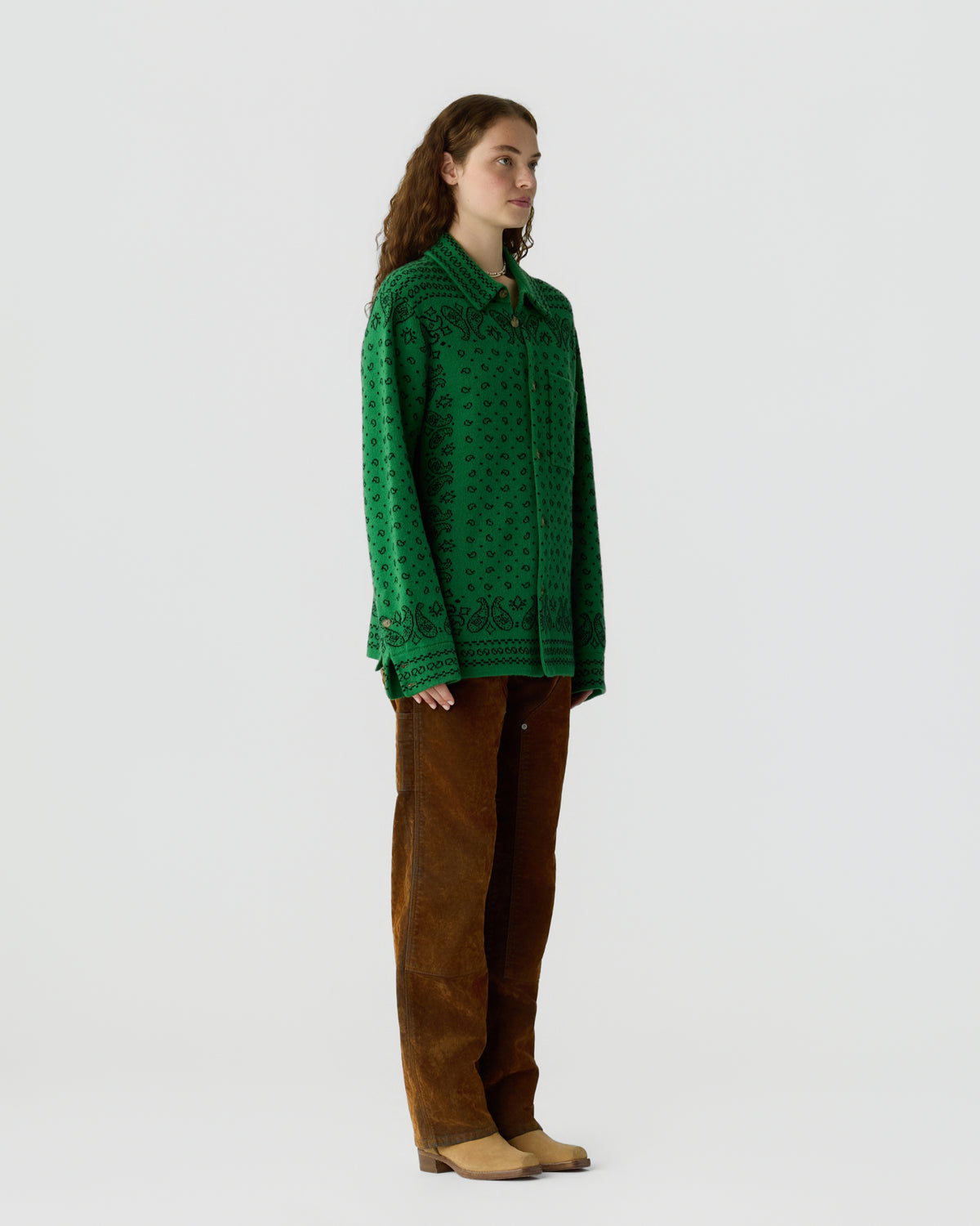 Woman with long curly hair wearing a green patterned button-up sweater, brown corduroy pants, and beige shoes standing against a plain light background in profile view.