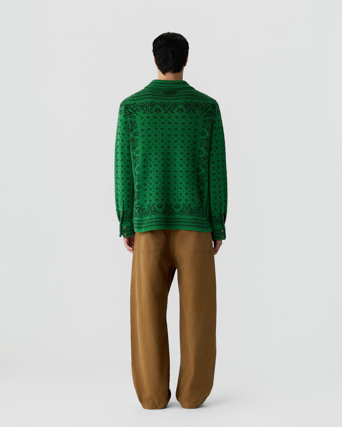 Man wearing a green long-sleeve shirt with a black paisley pattern and loose brown pants standing against a plain light gray background, viewed from the back.