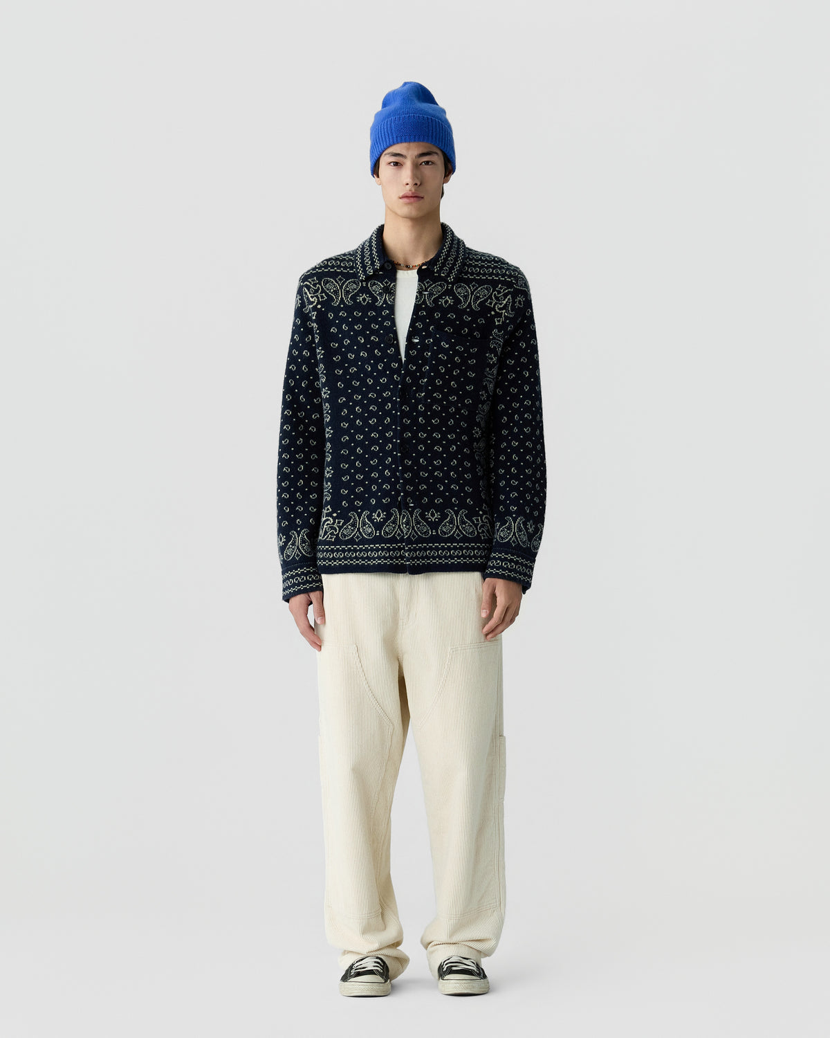 Young man wearing a blue beanie, a dark patterned button-up cardigan, cream wide-leg pants, and black and white sneakers standing against a plain light gray background.