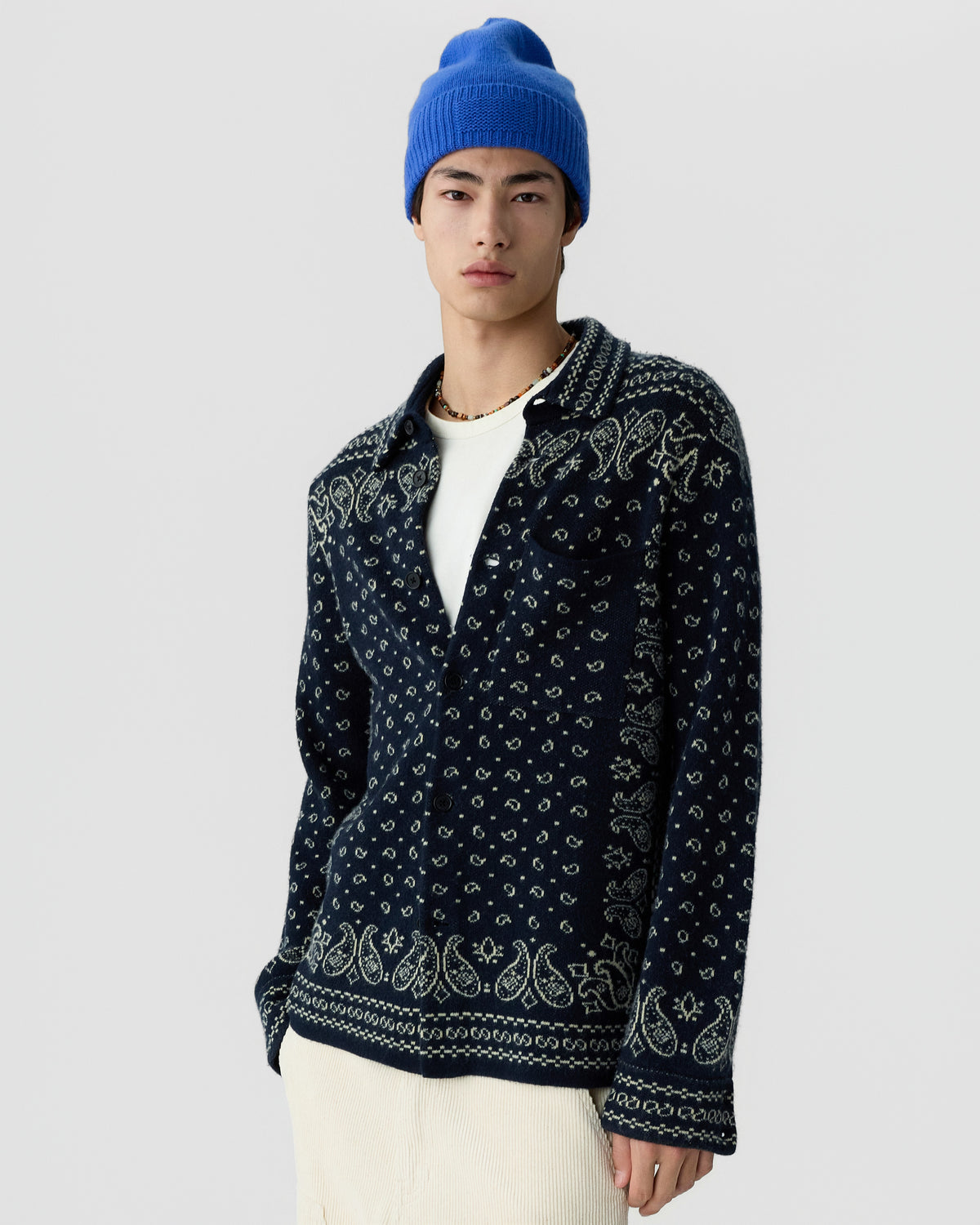 A young man wearing a blue knit beanie and a dark blue patterned button-up cardigan over a white shirt, standing against a plain light gray background.