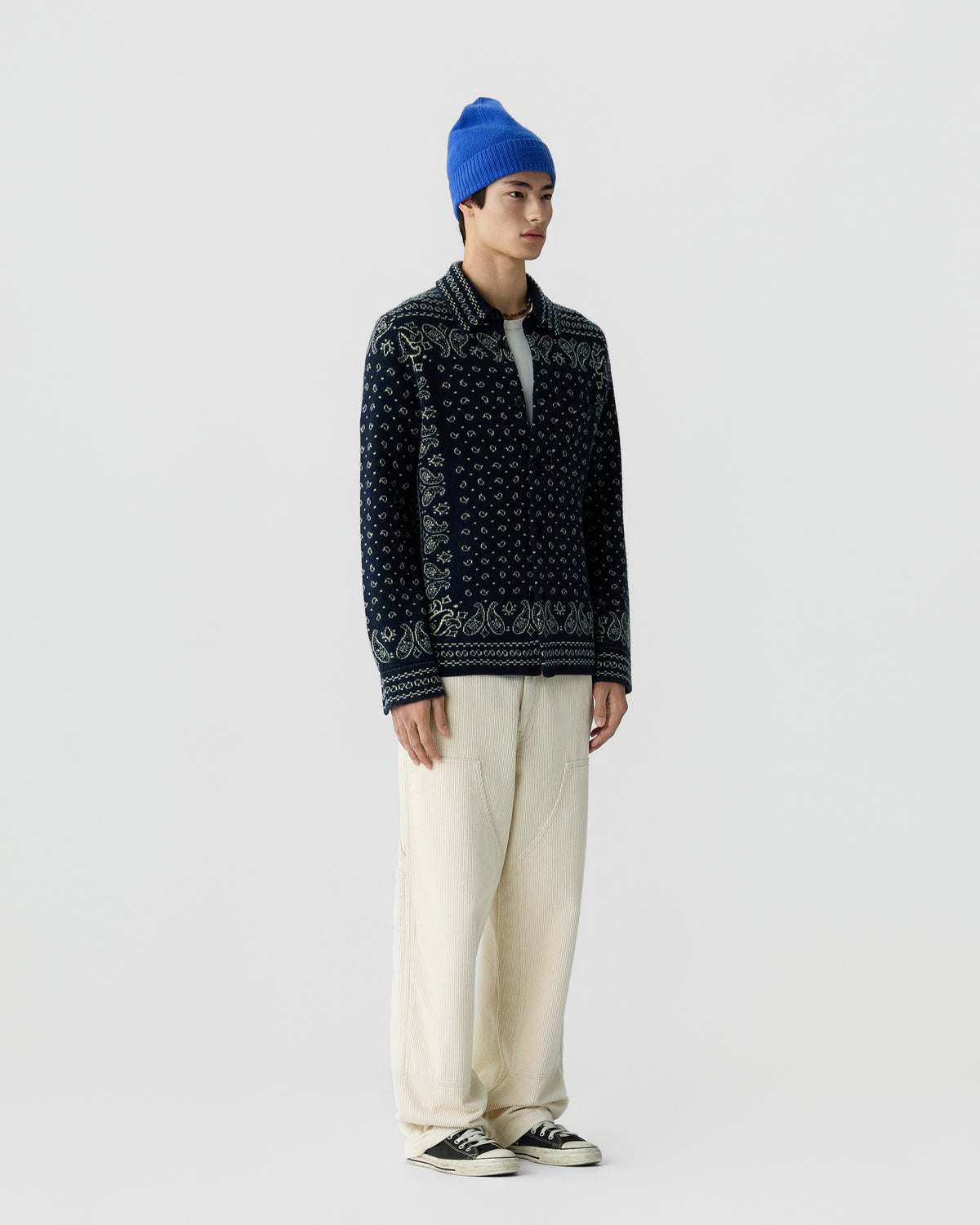 Young man wearing a blue knit beanie, dark patterned cardigan over a white shirt, loose cream-colored pants, and black sneakers standing against a plain light background.
