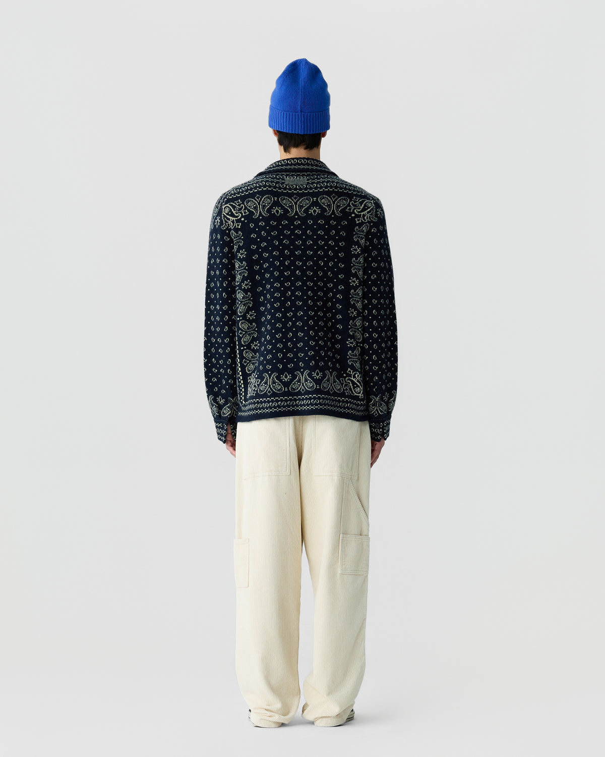 Person wearing a blue beanie, a dark patterned sweater with intricate designs on the back, and loose off-white pants, standing facing away against a plain light background.